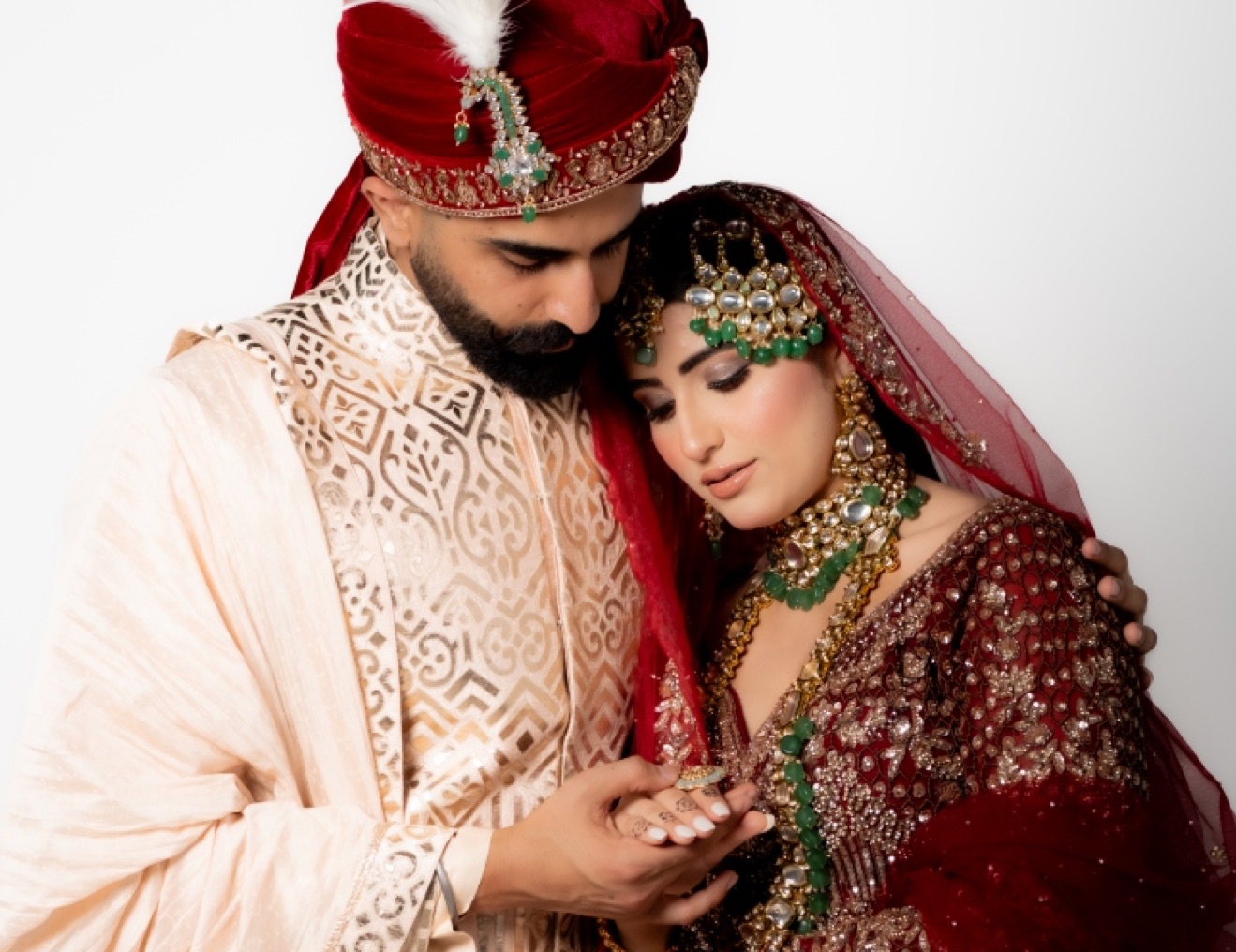 Groom in an ivory sherwani and red turban holding his bride, who wears a red lehenga and emerald jewellery