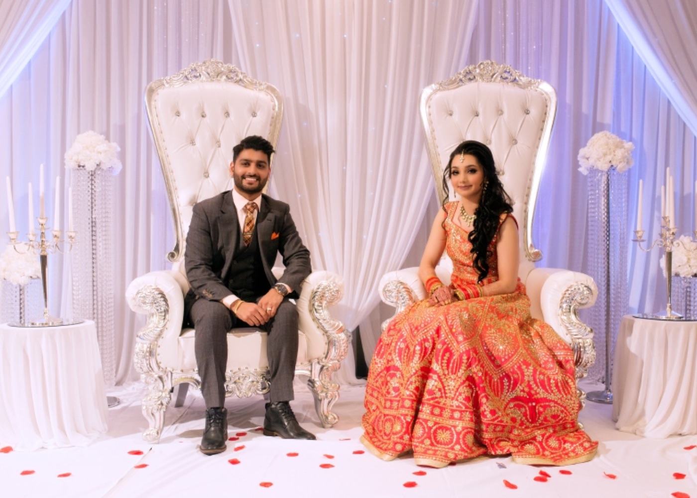 Couple seated on white thrones on a draped stage scattered with petals