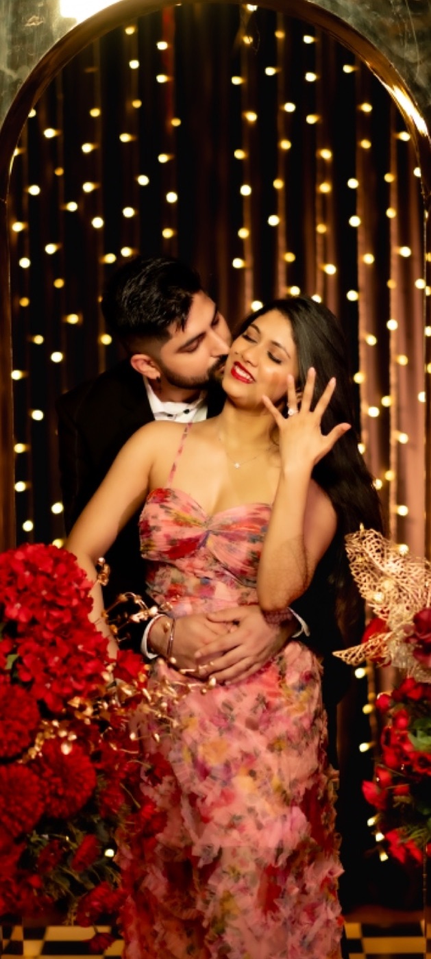 Woman showing her engagement ring as her partner kisses her cheek under a lit arch